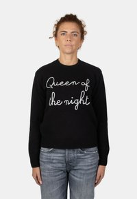 Maglione nero con la scritta "Regina della notte" ricamata in bianco. Collo e polsini a coste, abbinato a jeans azzurri chiari.
