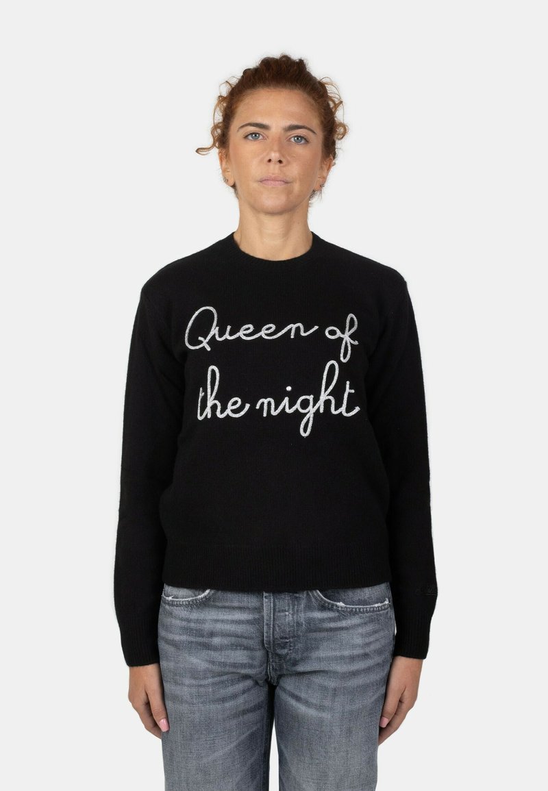 Maglione nero con la scritta "Regina della notte" ricamata in bianco. Collo e polsini a coste, abbinato a jeans azzurri chiari.