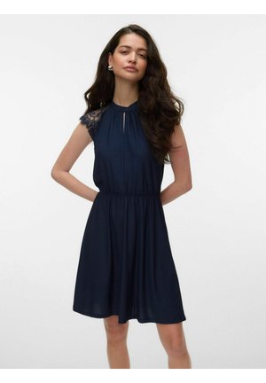 Femme aux longs cheveux foncés portant une robe sans manches bleu marine avec des épaules en dentelle, debout devant un fond clair uni.