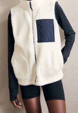 Persoon draagt een wit fleecevest met een zwarte borstzak met rits over een zwart shirt met lange mouwen en zwarte korte broek, hand rustend op de heup.