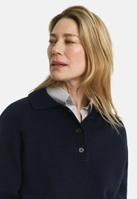 Maglione blu navy con colletto a coste, apertura con tre bottoni e tessuto strutturato. Indossato sopra una camicia a righe chiara con colletto.