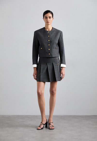 Grijs geruite cropped blazer met gouden knopen en witte manchetjes, gecombineerd met een bijpassende geplisseerde minirok met zijgespen. Zwarte strappy hakken.