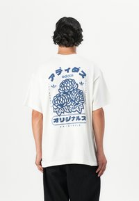 White cotton t-shirt featuring a blue floral design with Japanese text and Adidas branding on the back. Loose fit with short sleeves.