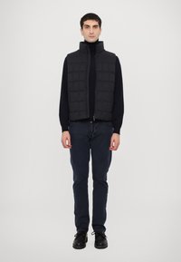 Black puffer vest with high collar, featuring a quilted design. Worn over a black turtleneck and paired with dark blue pants and black shoes.