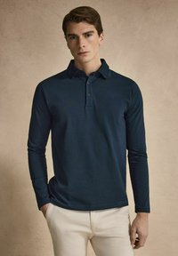 Långärmad marinblå pikétröja med mjukt tyg, treknapparsförslutning och klassisk krage, tillsammans med ljusa byxor.