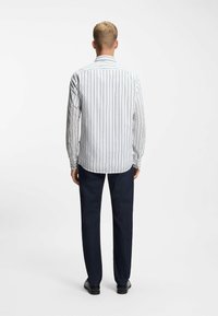 Wit overhemd met lange mouwen met verticale blauwe strepen, een afgeronde zoom en knoopmanchetten, gecombineerd met donkerblauwe broek en zwarte schoenen.