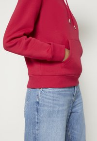 Czerwona bluza z kapturem, z przednią kieszenią, kapturem ze sznurkiem oraz ściągaczami na mankietach i u dołu. W zestawie z jasnoniebieskimi dżinsami. Miękka tekstura materiału.