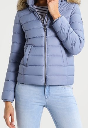 Hellblaue gesteppte Steppjacke mit pelzbesetzter Kapuze, getragen über einem gestreiften Hemd, kombiniert mit hellblauen Denim-Jeans.