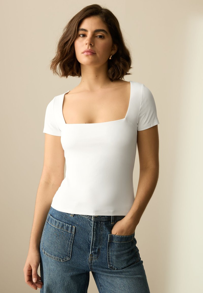 T-shirt blanc ajusté avec un col carré et des manches courtes, associé à un jean en denim bleu taille haute avec poches arrière.