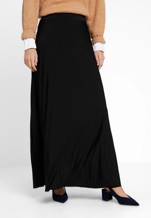 Femme portant une longue jupe noire, un pull marron clair par-dessus une chemise blanche, et des chaussures bleu marine à talons bas et bouts pointus sur fond blanc.