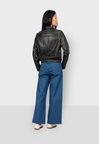 Černá kožená zkrácená bunda s vysokým límcem a elastickým lemem, doplněná širokými modrými džínovými kalhotami a světlými botami.