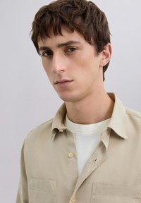 Camisa beige de botones con cuello, dos bolsillos en el pecho y textura suave, superpuesta sobre una camiseta blanca. Fondo liso.