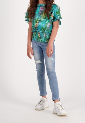 Pige iført en teal farvet blomstret top med flæseærmer, lyse, revnede skinny jeans og hvide chunky sneakers, der står på en ensfarvet baggrund.