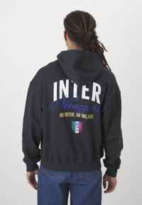 Sweatshirt preto com "INTER" em letras brancas grandes nas costas, com detalhes em azul em letra cursiva e um emblema colorido perto da bainha.