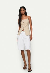 Beige knapplös västtop med fyrkantig halsringning, matchad med vita knälånga shorts och svarta flip-flops. Slät tyg och minimalistisk design.