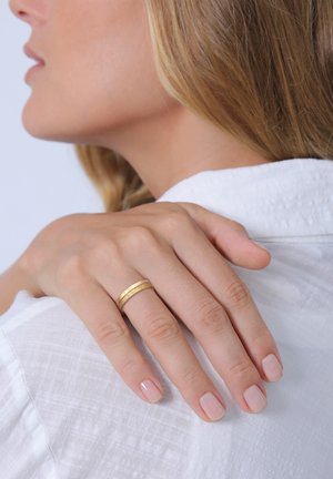 Gold ring with a textured band worn on the hand, resting on a white, wrinkled fabric. Nails are manicured with a light pink polish.