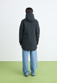 Casaco impermeável preto com capuz, apresentando uma cintura ajustada, mangas longas e um design de comprimento médio, combinado com calças jeans leves de cor azul clara.