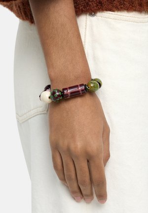 Nadgarstek z bransoletką z koralików z dużymi zielonymi, fioletowymi i kremowymi koralikami noszony na białych spodniach i brązowym, teksturowanym rękawie swetra.