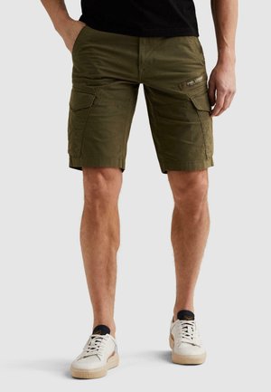 Mężczyzna w oliwkowych spodenkach cargo z wieloma kieszeniami, czarnej koszulce i białych trampkach, stoi z jedną ręką w kieszeni.