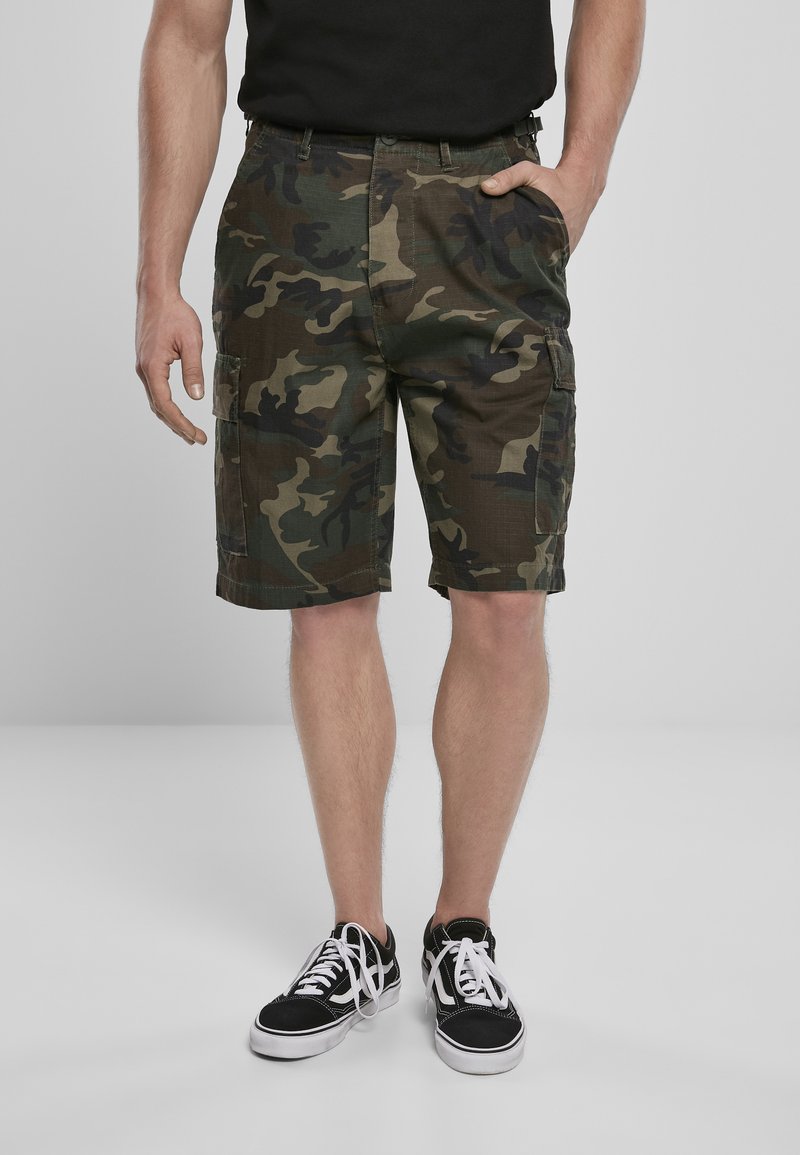 Zöld és barna árnyalatú, szövetből készült kamuflázs cargo rövidnadrág, cargo zsebekkel. Fekete sneaker cipőkkel párosítva, fehér fűzőkkel.