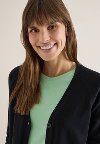 Femme souriante aux longs cheveux bruns portant un haut vert menthe et un cardigan noir boutonné, sur un fond clair uni.