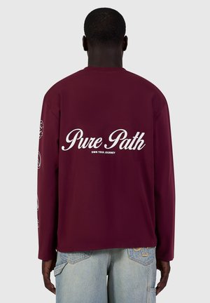 Person wearing a maroon long-sleeve shirt with white text "Pure Path Own Your Journey" on the back and light blue denim jeans.