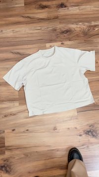 Hvid, kortærmet t-shirt lagt fladt på et trægulv; har en simpel rund hals og en afslappet pasform med en glat tekstur.
