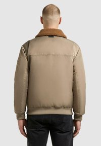 Mand iført en beige polstret jakke med brun fårskindskrave og sorte jeans, vist fra ryggen mod en ensfarvet lys baggrund.