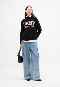 Czarny sweter dzianinowy z napisem "DKNY NEW YORK" w różowym kolorze, nałożony na jasnoróżową koszulę, w zestawieniu z szerokimi niebieskimi dżinsami i czarnymi butami.
