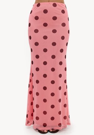 Longue jupe rose avec de gros pois rouge foncé portée sur les pieds, montrée de la taille vers le bas sur un fond blanc.