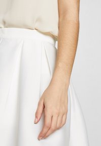 Gonna bianca a pieghe con una vita aderente, abbinata a un top beige chiaro. Primo piano di una mano appoggiata sul tessuto della gonna.