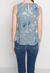 Hihaton paita sinisestä kankaasta, jossa on vaaleanpunaisia, keltaisia ja tummansinisia kukkakuoseja. Takana on nappikiinnitys ja avonainen yksityiskohta.