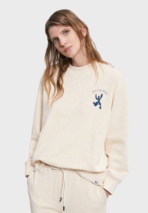 Giovane donna con capelli castano chiaro lunghi, indossa una felpa crema e pantaloni abbinati, con le mani nelle tasche, su uno sfondo semplice grigio chiaro.