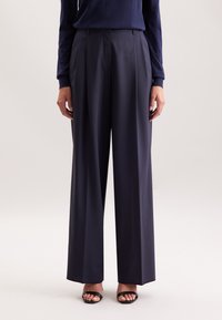 Persoon in een marineblauwe wijde broek, een marineblauw lange mouw top en zwarte open-teenschoenen, staand tegen een effen lichte achtergrond.