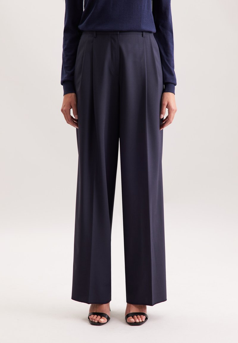 Persoon in een marineblauwe wijde broek, een marineblauw lange mouw top en zwarte open-teenschoenen, staand tegen een effen lichte achtergrond.