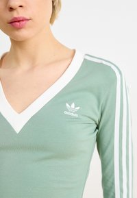 Zelena dolga majica z V-izrezom, belim robom, tremi belimi črtami na rokavih in majhnim belim Adidasovim logotipom na prsih.