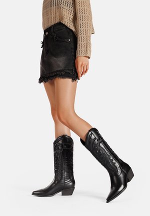 Stivali da cowboy neri con punta affusolata, design in rilievo, tacco basso e pelle texturizzata; abbinati a una gonna di jeans nera sfilacciata.