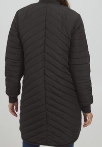 Femme portant une longue veste noire matelassée avec des coutures en chevrons et horizontales, debout les bras détendus devant un fond clair uni.