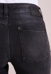 Vaqueros de mezclilla gris oscuro con un diseño ajustado. La parte posterior cuenta con un parche de cuero negro texturizado y bolsillos estructurados con detalles de costura.