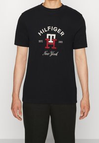 Černé bavlněné tričko s krátkým rukávem; na přední straně je vyšívané logo "HILFIGER", "1985" a "New York" s červenými a bílými akcenty.