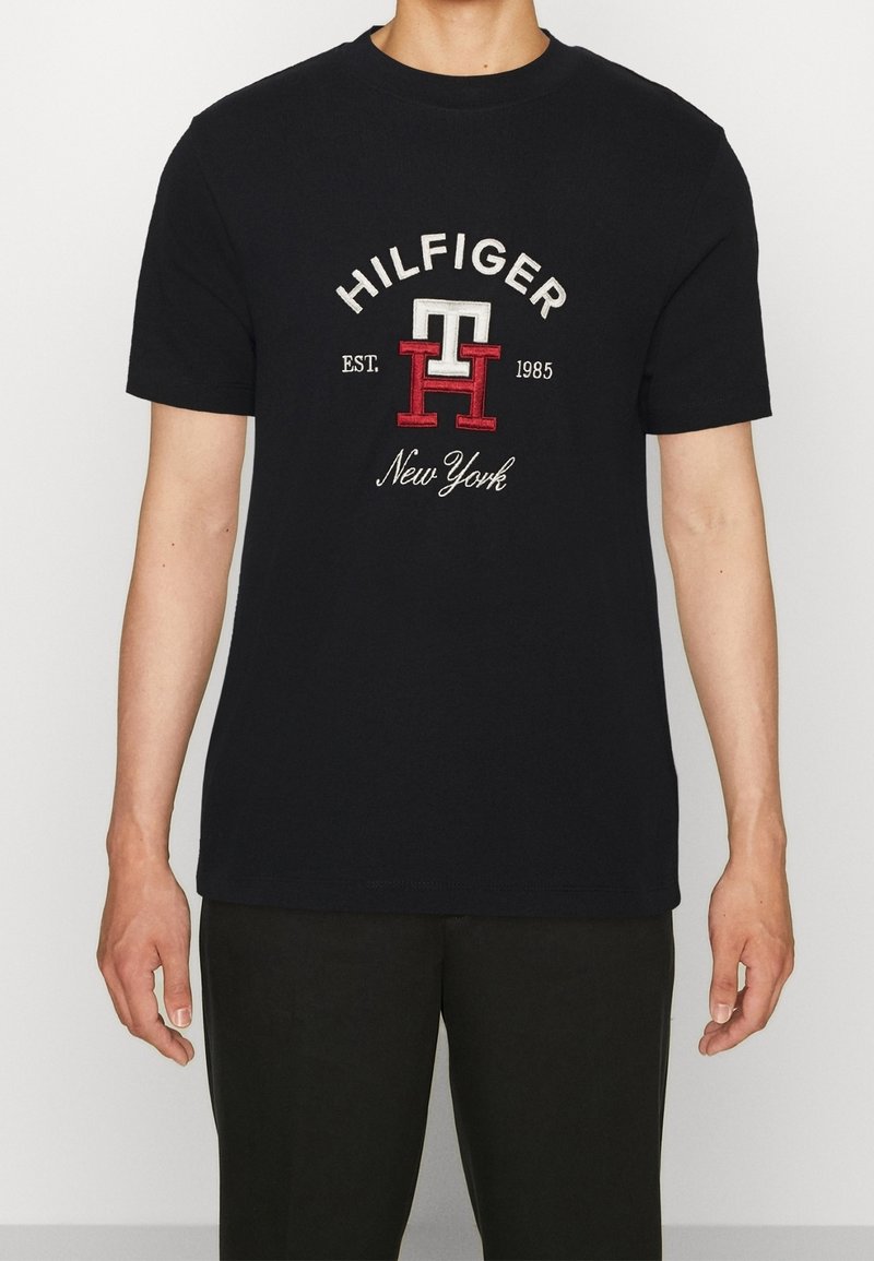 Černé bavlněné tričko s krátkým rukávem; na přední straně je vyšívané logo "HILFIGER", "1985" a "New York" s červenými a bílými akcenty.