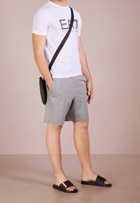 Biała koszulka z nadrukowanym logo, szare spodenki, czarne klapki z czerwonymi akcentami i czarna torba na ramię. Prosty, casualowy strój.