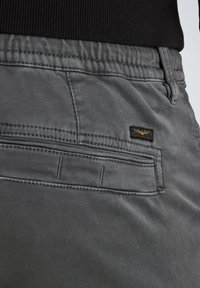 Graue Stoffhosen mit glatter Textur, ausgestattet mit einer hinteren Tasche und einem kleinen gestickten Logo. Der Bund ist elastisch für zusätzlichen Komfort.