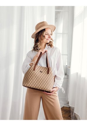 Beige Handtasche mit Muster und braunen Ledergriffen, auf der Vorderseite mit einem Logo versehen. Das Modell trägt ein weißes Hemd und beige Hosen.