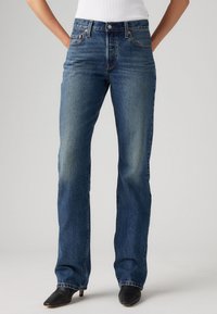 Blauwe denim jeans met een rechte pasvorm, middelmatige wassing, subtiele vervaging, vijf zakken ontwerp en koperen knopenaccenten. Ggedragen met zwarte hakken.
