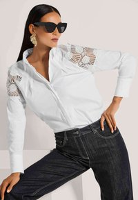 Witte button-up blouse met gedetailleerde kantaccenten op de schouders, gecombineerd met donkere spijkerbroek. Model draagt zwarte zonnebril.