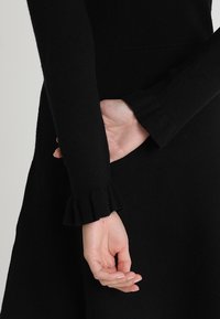 Abito nero con maniche lunghe e polsini a costine. Il tessuto appare morbido e texture, con una silhouette aderente e senza motivi visibili.