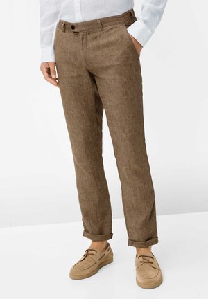 Uomo con pantaloni di lino marroni arrotolati, scarpe da barca beige in suede e camicia bianca a maniche lunghe, in piedi con una mano in tasca.