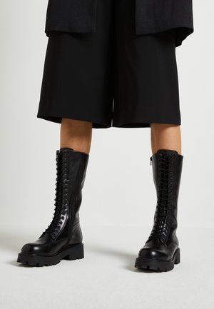 Osoba nosząca czarne, wysokie do kolan kozaki sznurowane z grubą podeszwą oraz czarne szerokie szorty na tle gładkiego jasnego tła.