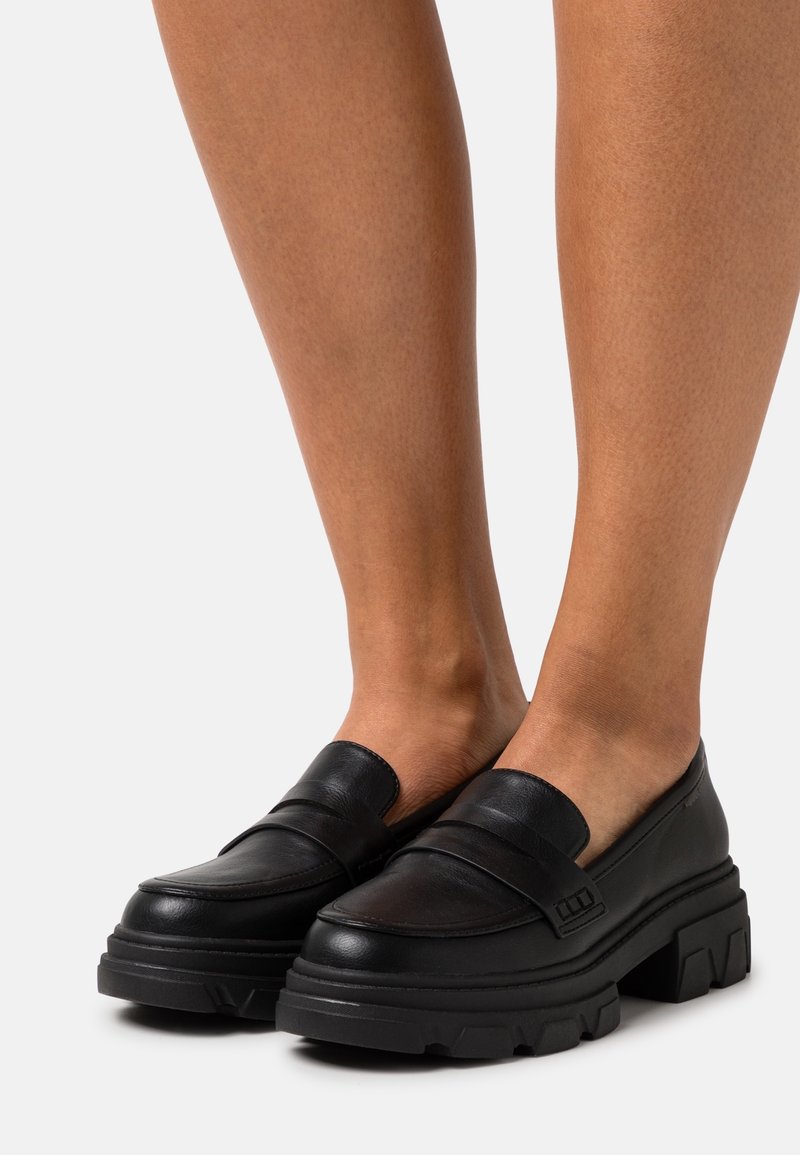 Mocassins noirs en cuir épais avec semelles épaisses et rainurées portés aux pieds sur un fond clair uni.